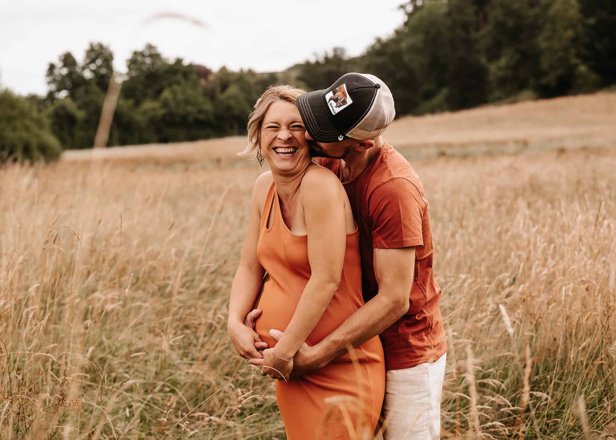 Pregnant woman in an orange dress stands in a sunlit field as her partner embraces her from behind and kisses her shoulder.