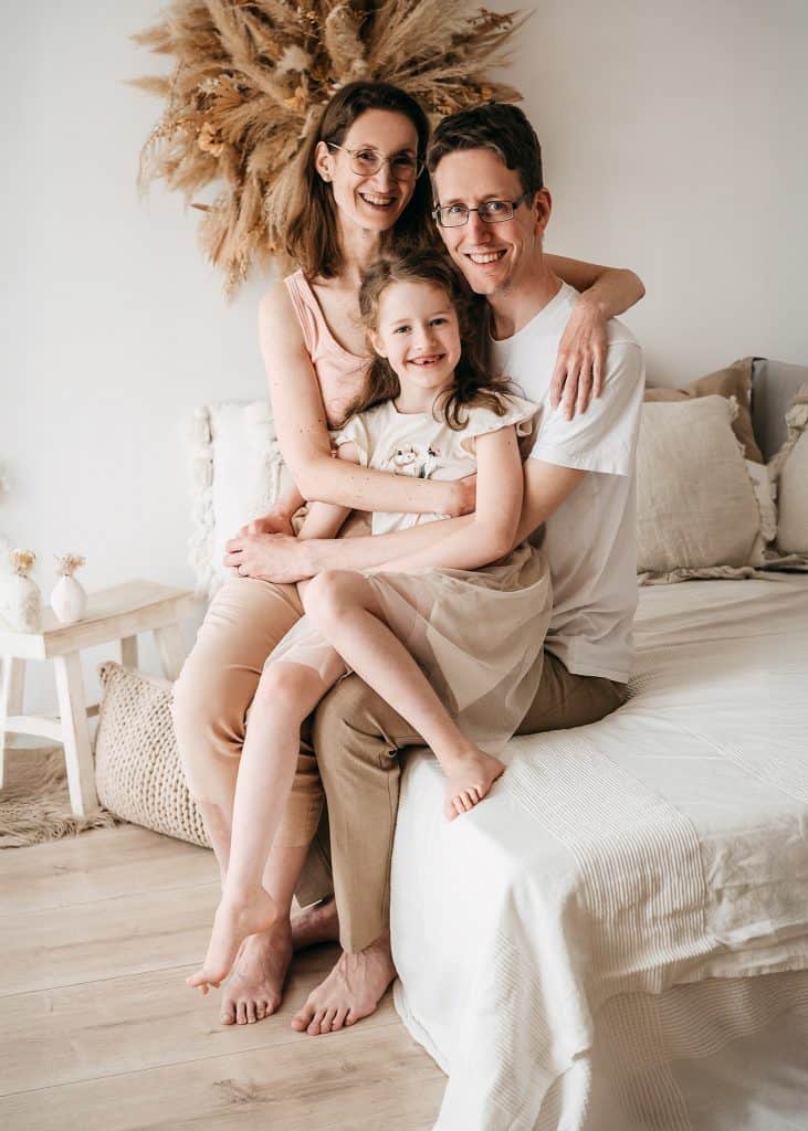Family of three—mother, father, and daughter—smiling and embracing on a bed in a bright, neutral-toned room.
