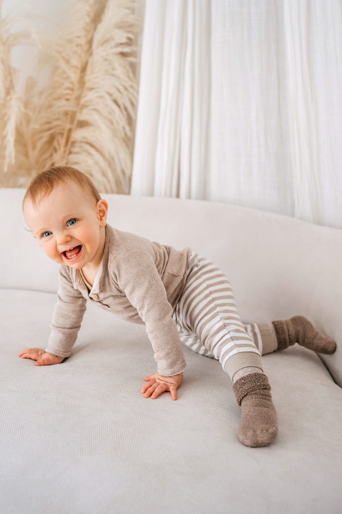 Smiling baby in a beige outfit crawling on a light sofa, looking at the camera with blue eyes.